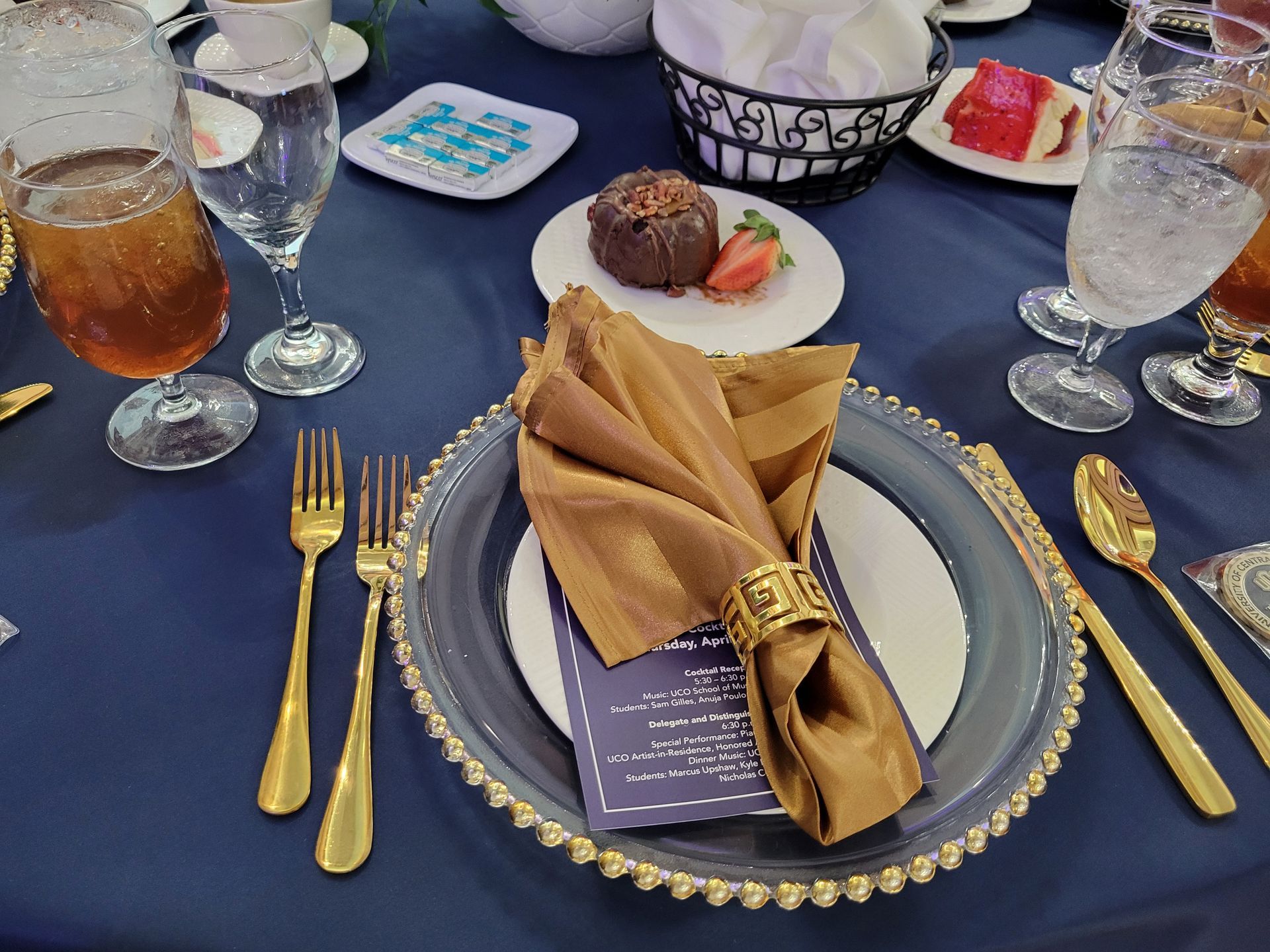 Formal table setting with gold silverware, navy tablecloth, gold napkin, and dessert.