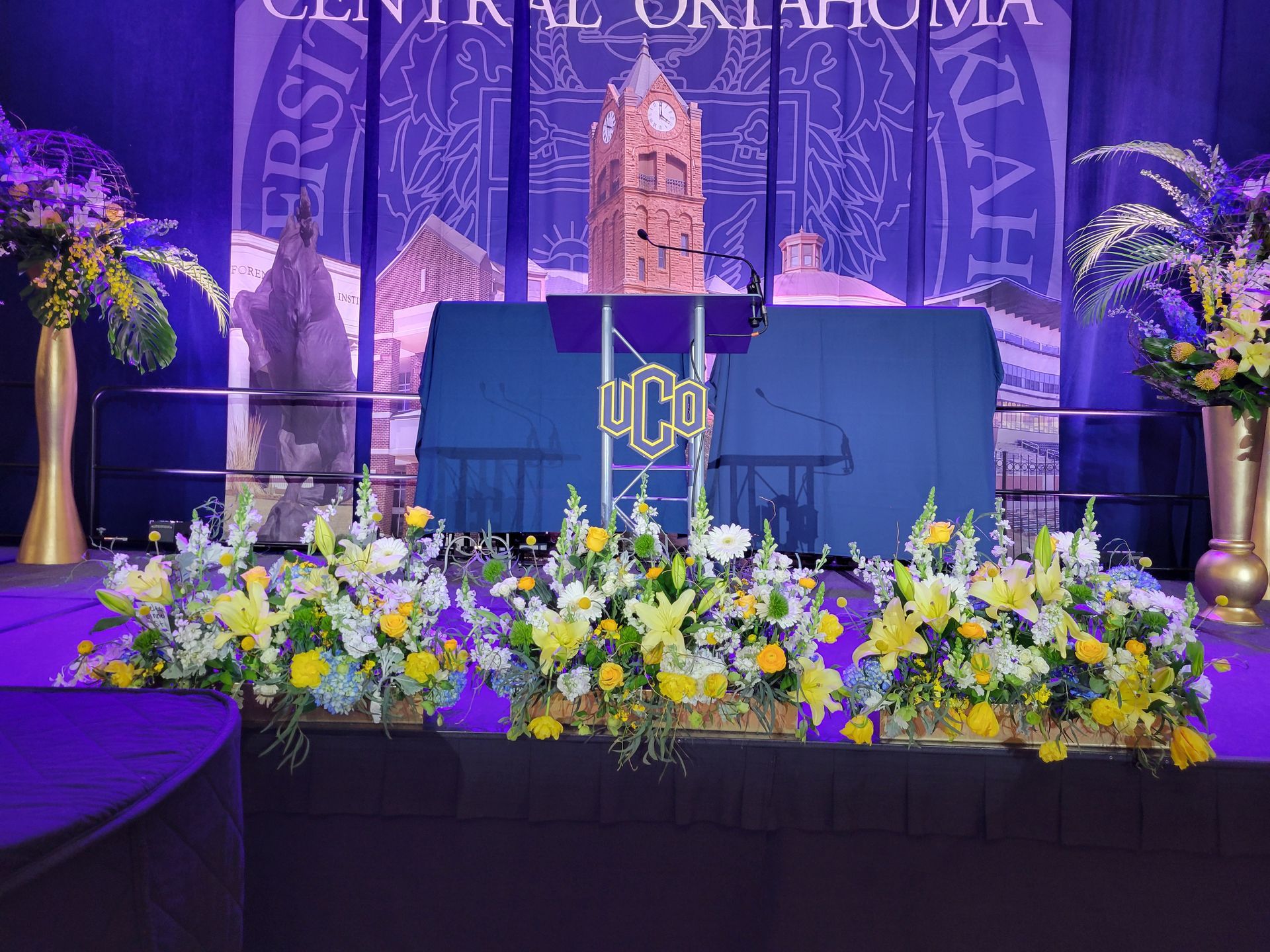 Stage decorated with yellow and white flowers for a university ceremony; backdrop of university logo.