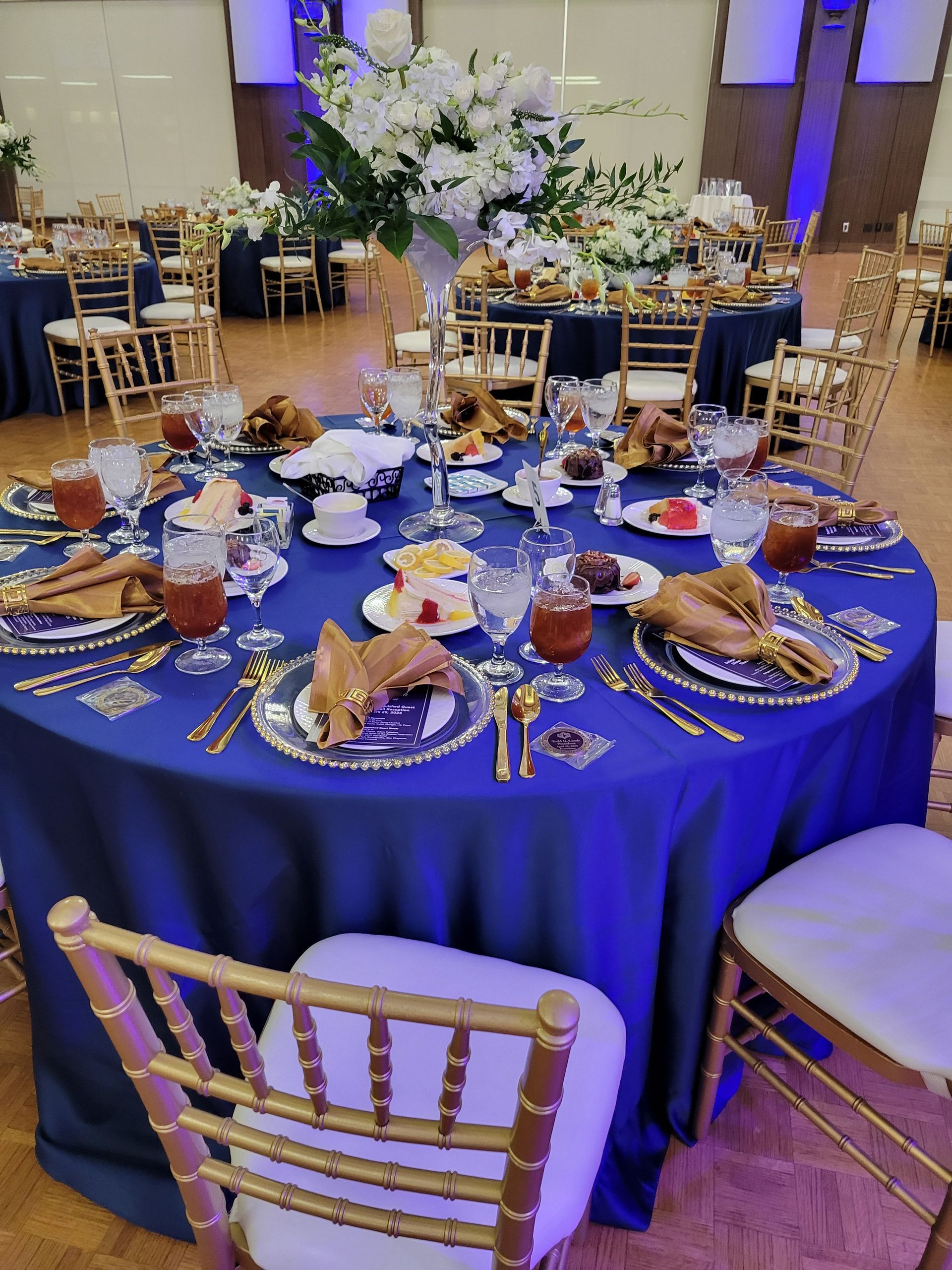 Round table set for a formal event with blue tablecloth, gold accents, floral centerpiece, and chairs.