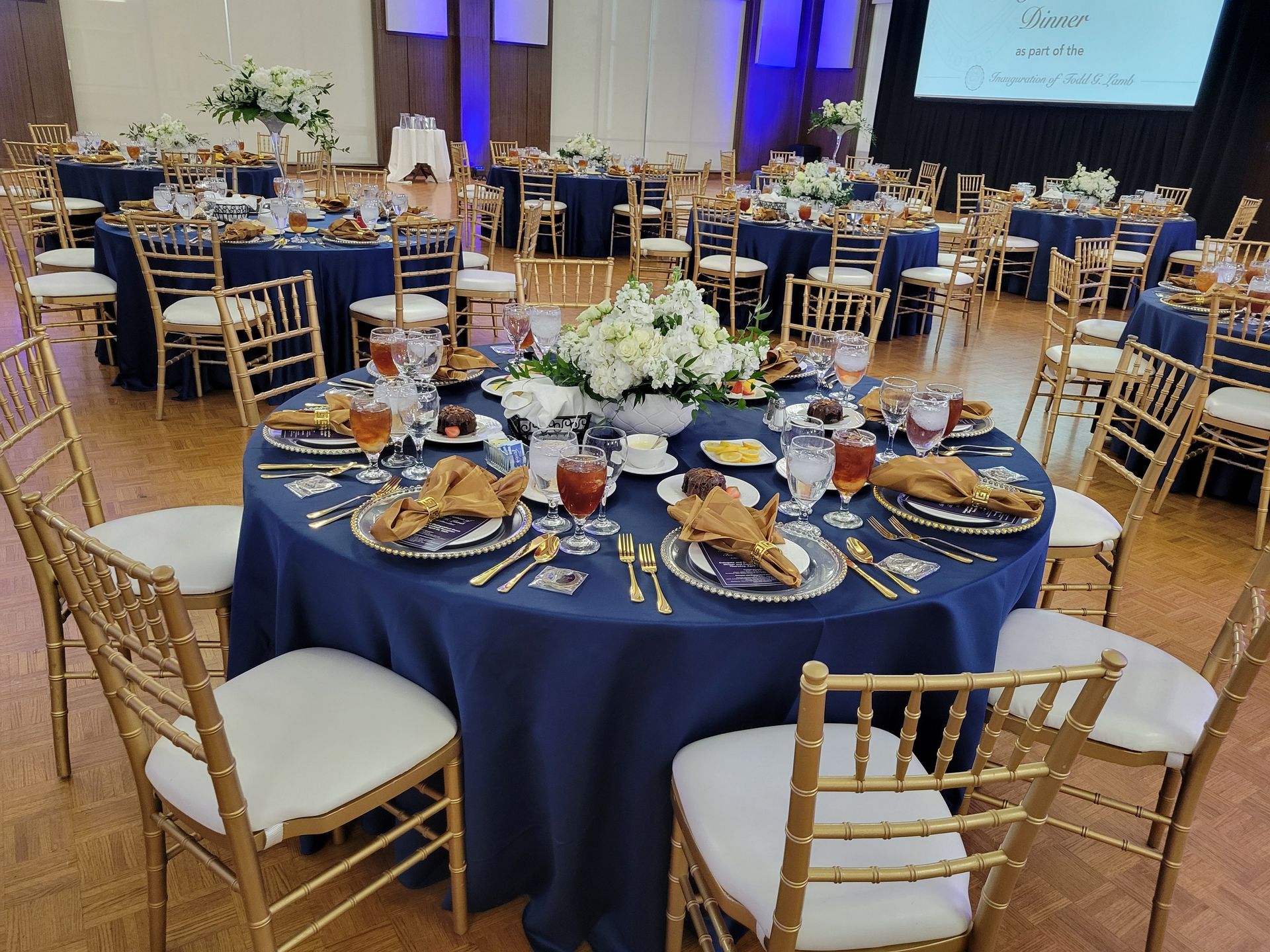 Reception hall with round tables set for a formal event. Navy blue tablecloths, gold chairs, white floral centerpieces.