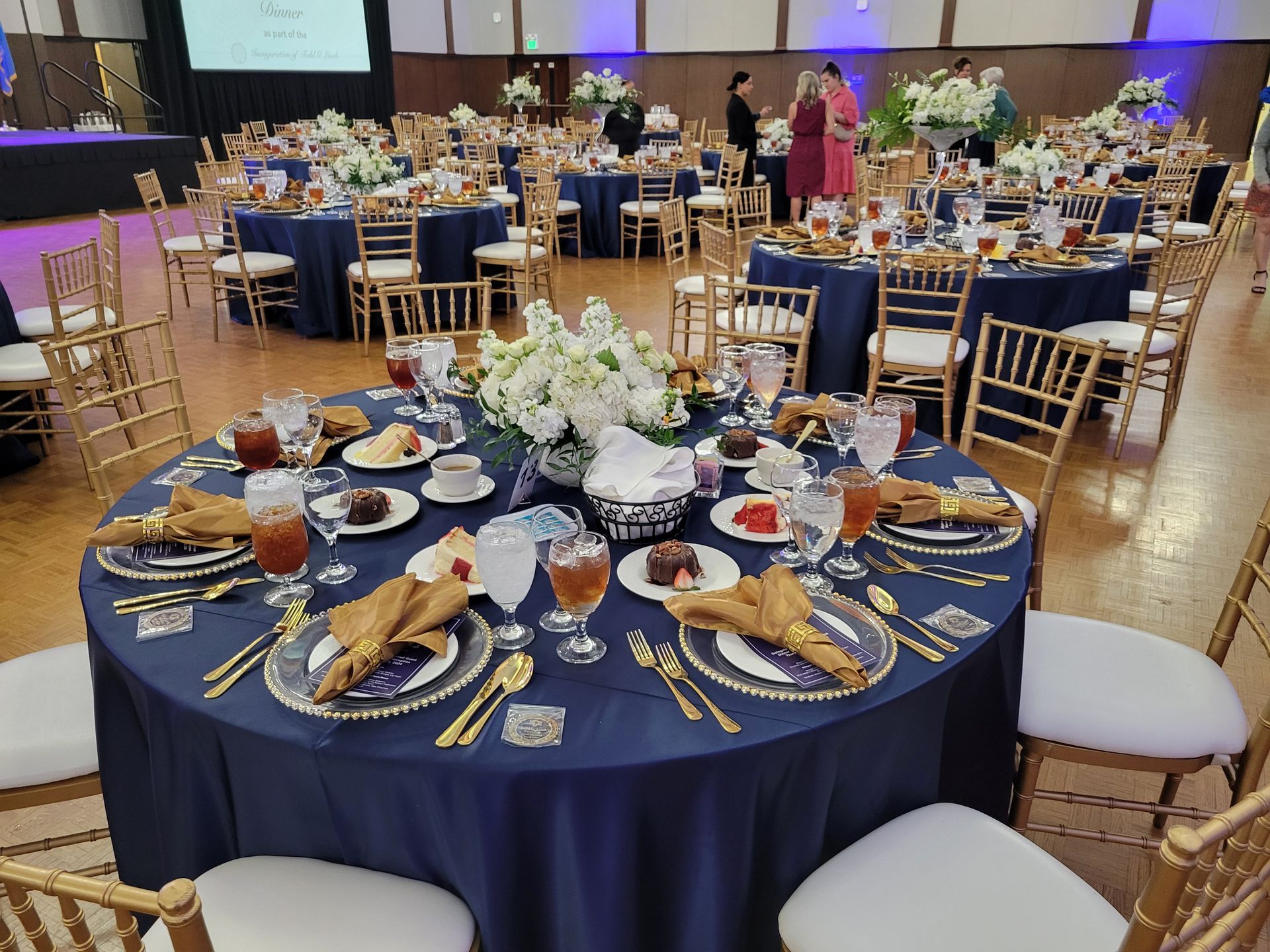 Round tables set for a formal event with navy tablecloths, gold chairs, and floral centerpieces.