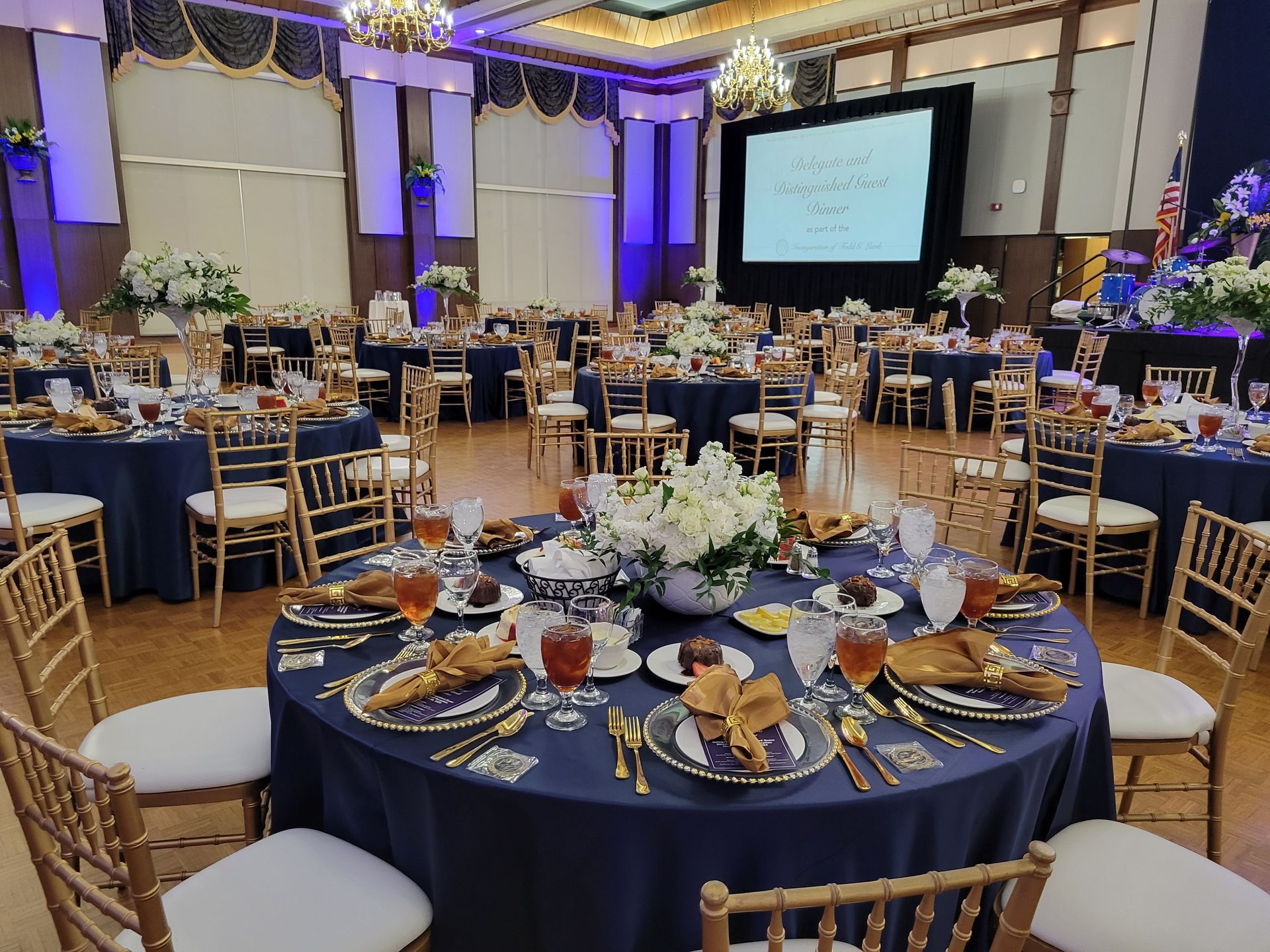 Formal event hall with round tables set for a meal. Dark blue tablecloths, gold chairs, and white flower centerpieces.