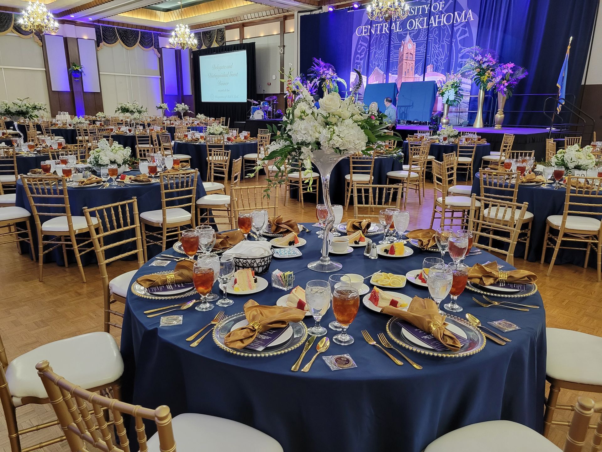 Formal event with round tables set for dinner. Navy blue tablecloths with gold chairs and floral centerpieces.