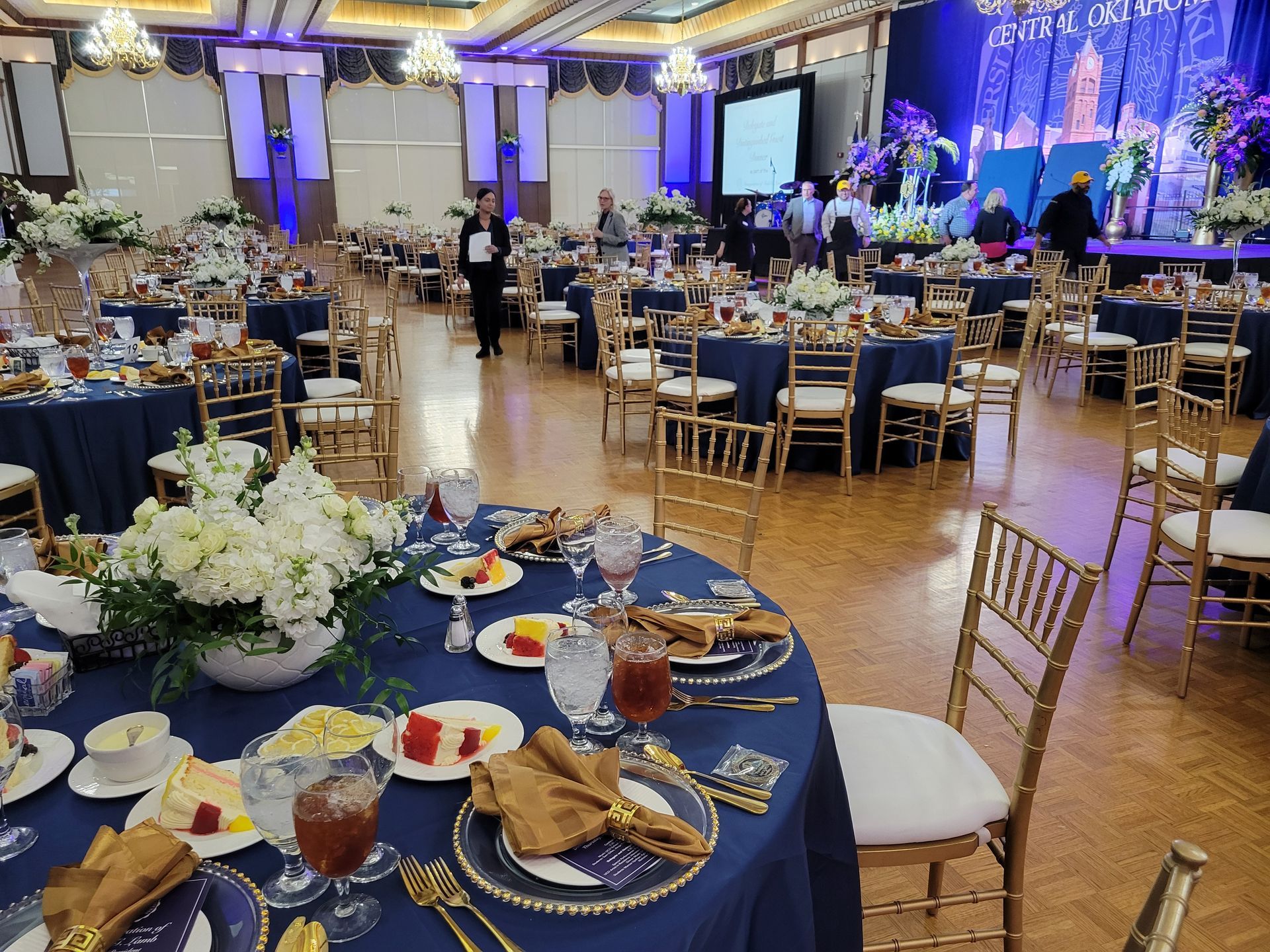 Banquet hall set with navy blue tablecloths, gold chairs, and floral centerpieces. People are mingling.
