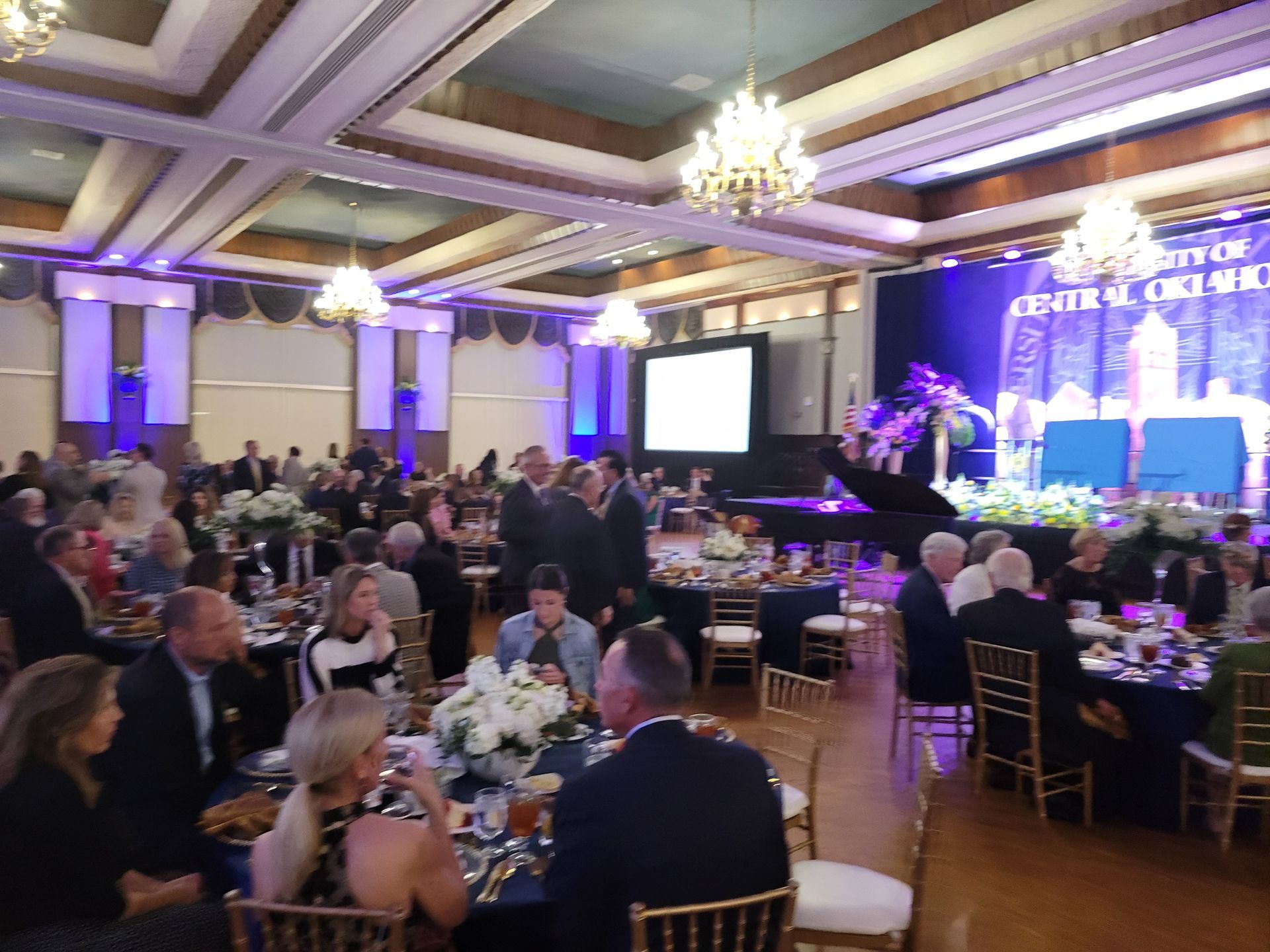 Formal event: People at tables in a large ballroom with stage, tables set, and a projection screen.