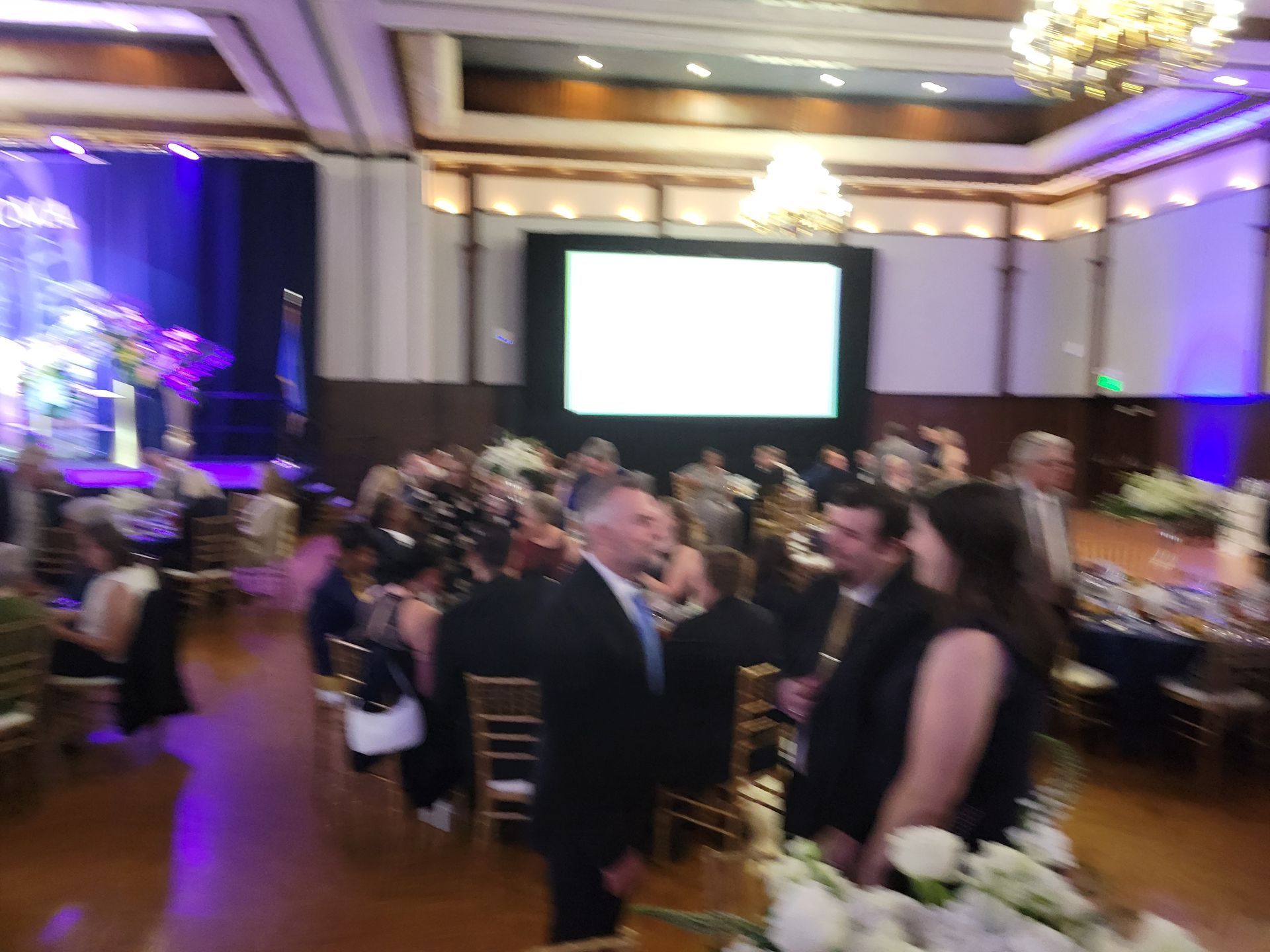 People at a formal event in a large ballroom with a screen, stage, and chandeliers.