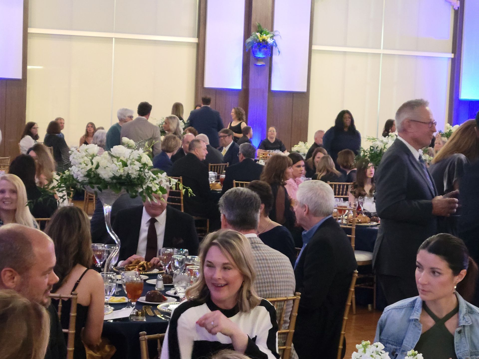 Large event with people at tables, many talking and some clapping. Blue and white decor.