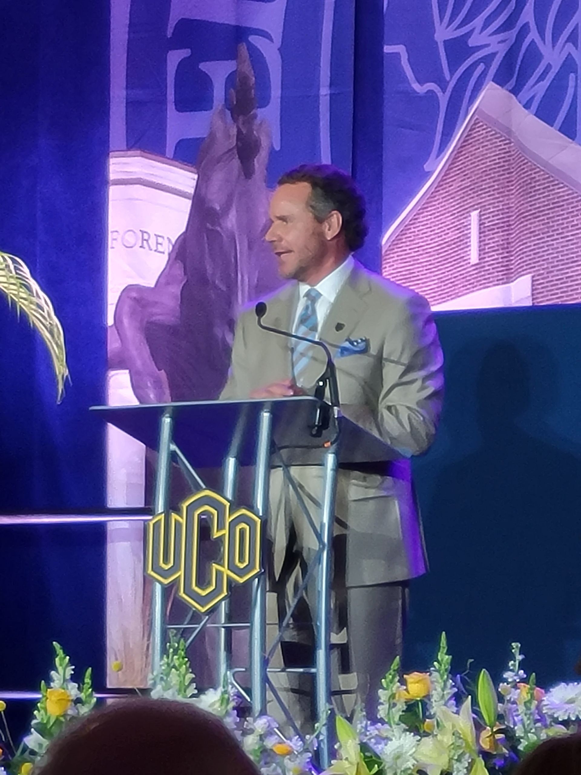 Man in suit speaks at a podium with UCO logo, flowers, and a statue background.
