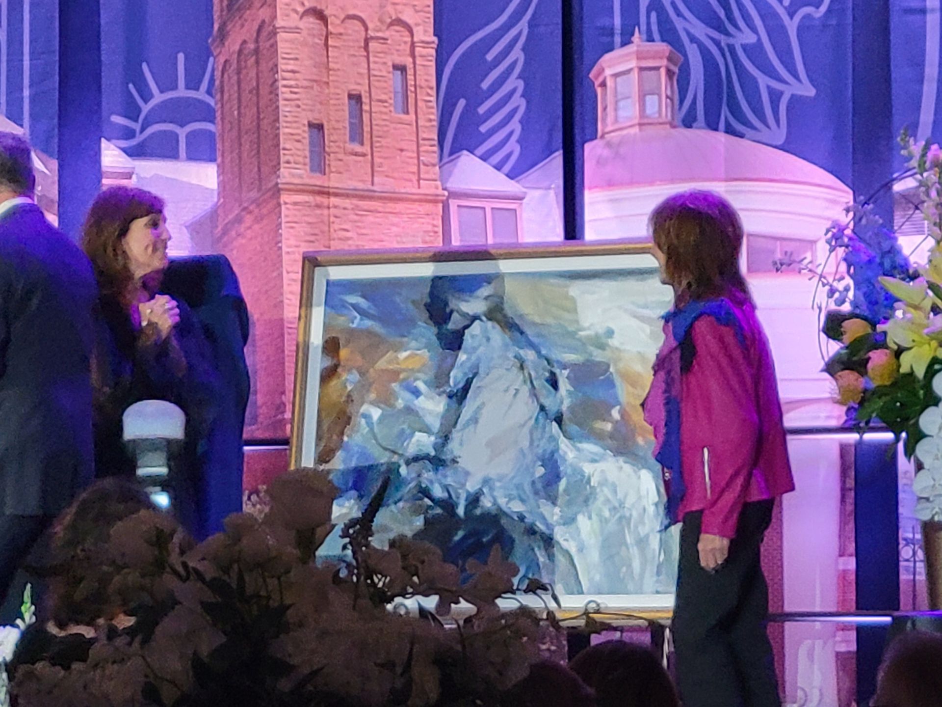 Two women beside a framed, abstract painting of mountains, indoors at a function.