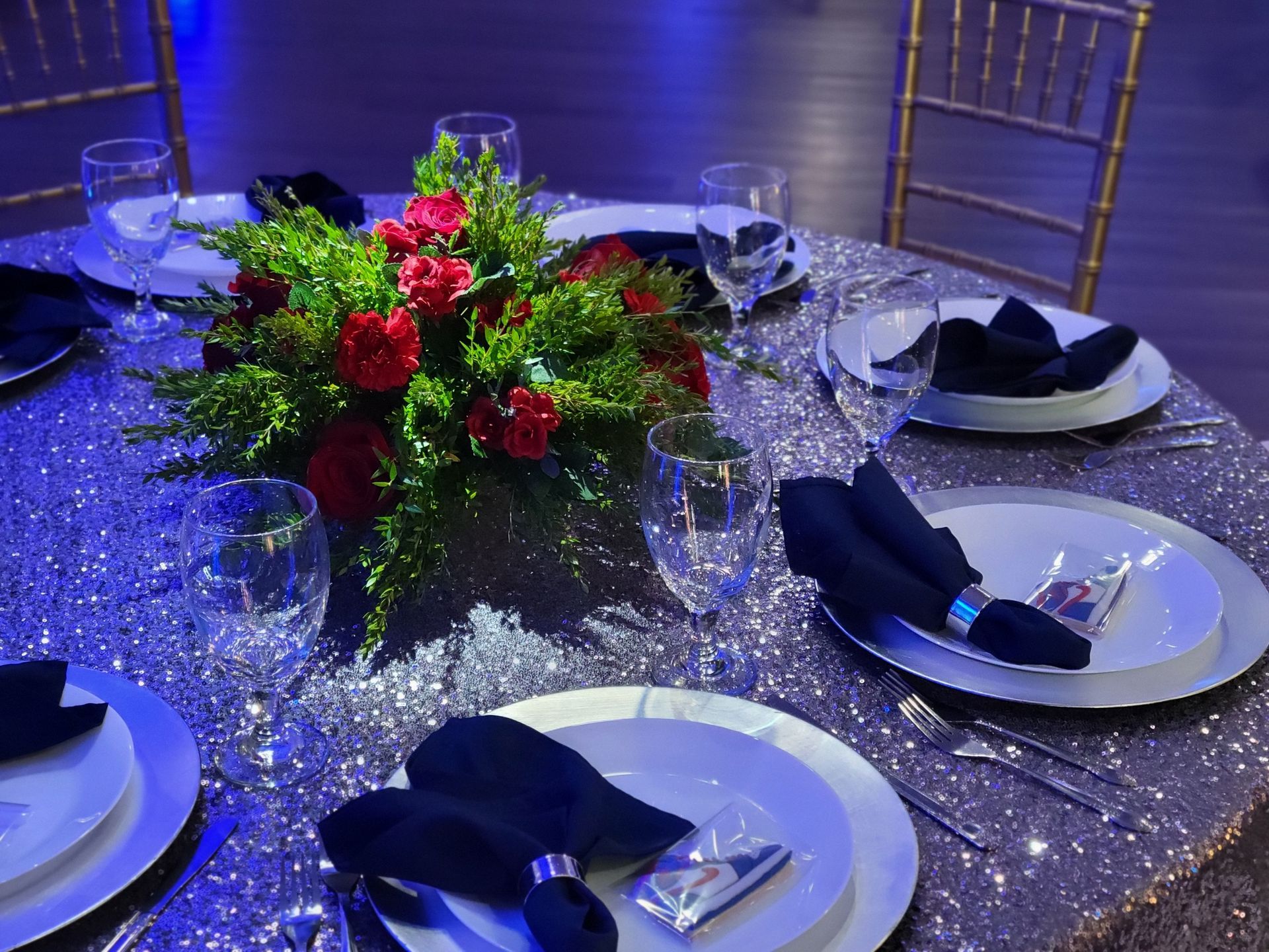 Elegant table setting with floral centerpiece on a sparkly silver tablecloth. Plates, silverware, and black napkins.