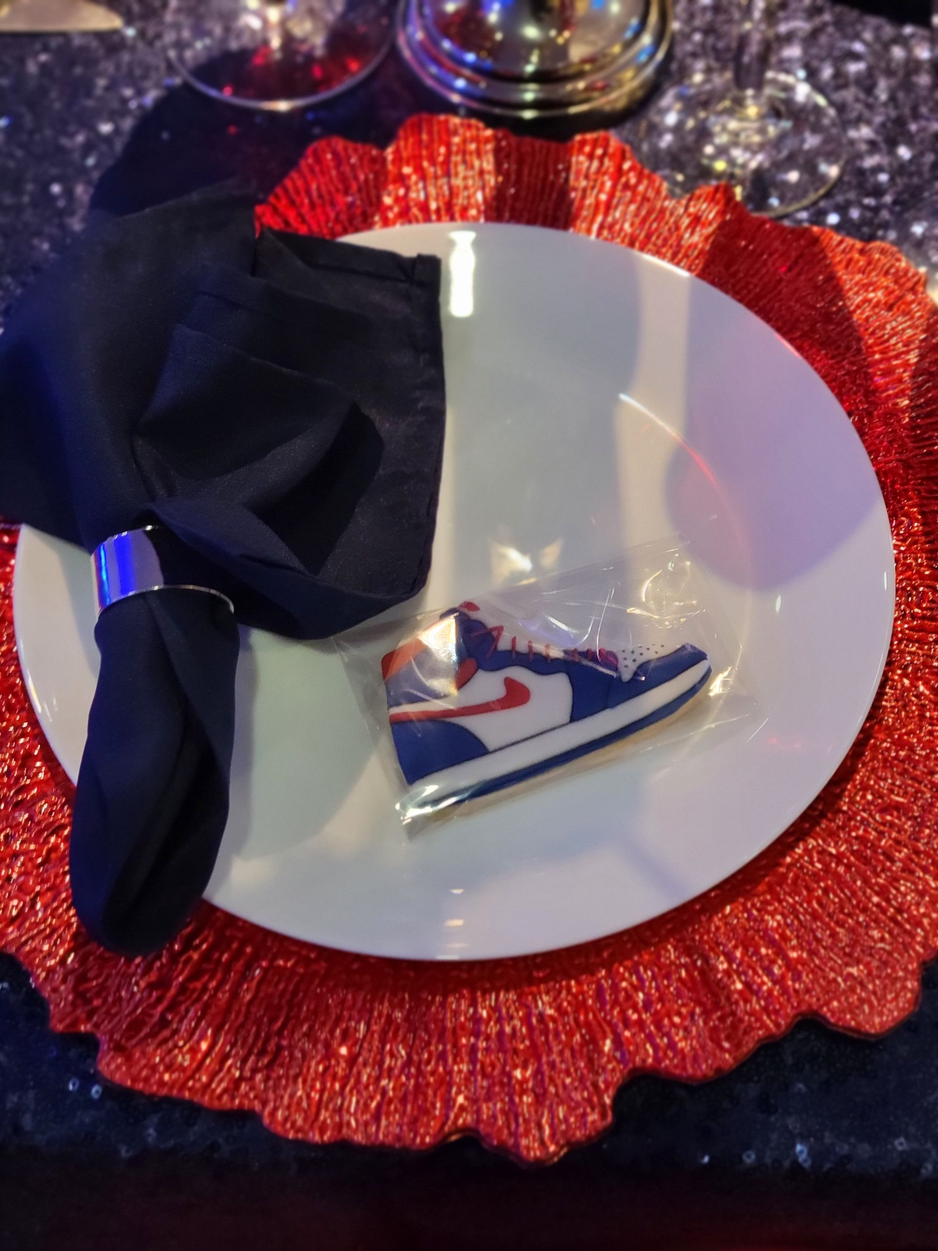 Place setting with red charger, white plate, black napkin, and sneaker-shaped treat.