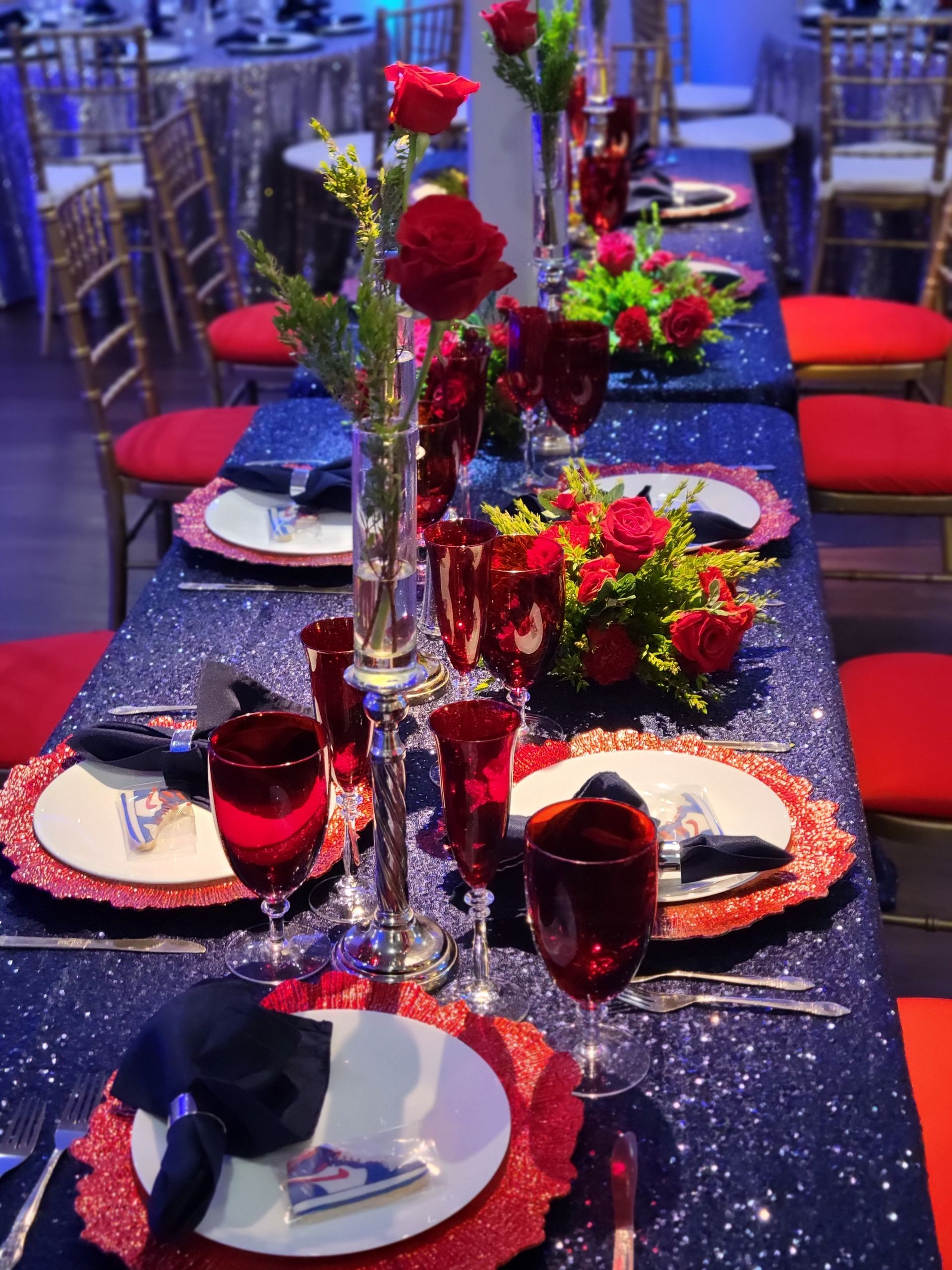 Elegant dining table set for event; navy sequin tablecloth, red roses, red placemats, white plates, and silver cutlery.