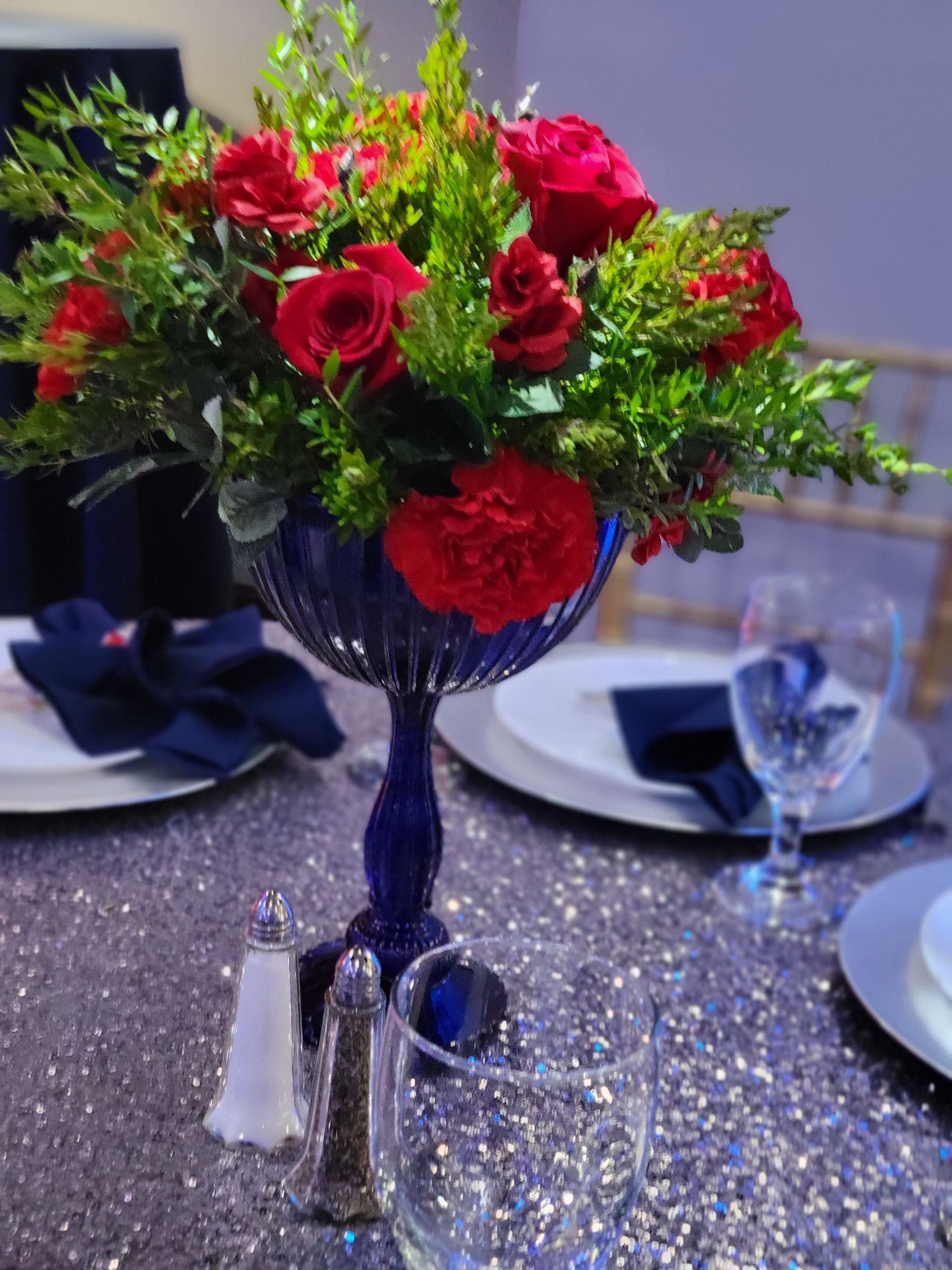 Centerpiece with red flowers in a blue vase on a sparkly table with place settings.