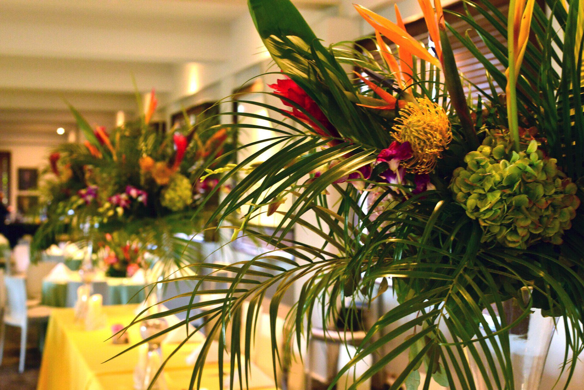 Tropical floral arrangements on tables at an event; yellow tablecloths and white chairs.