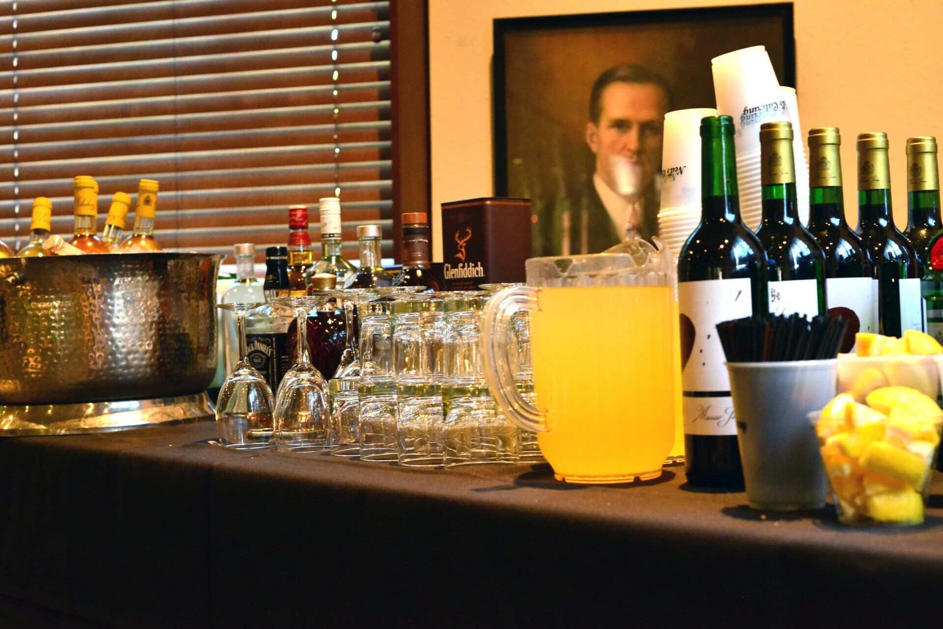 Bar with bottles of wine and liquor, glasses, a punch bowl, and a portrait in the background.