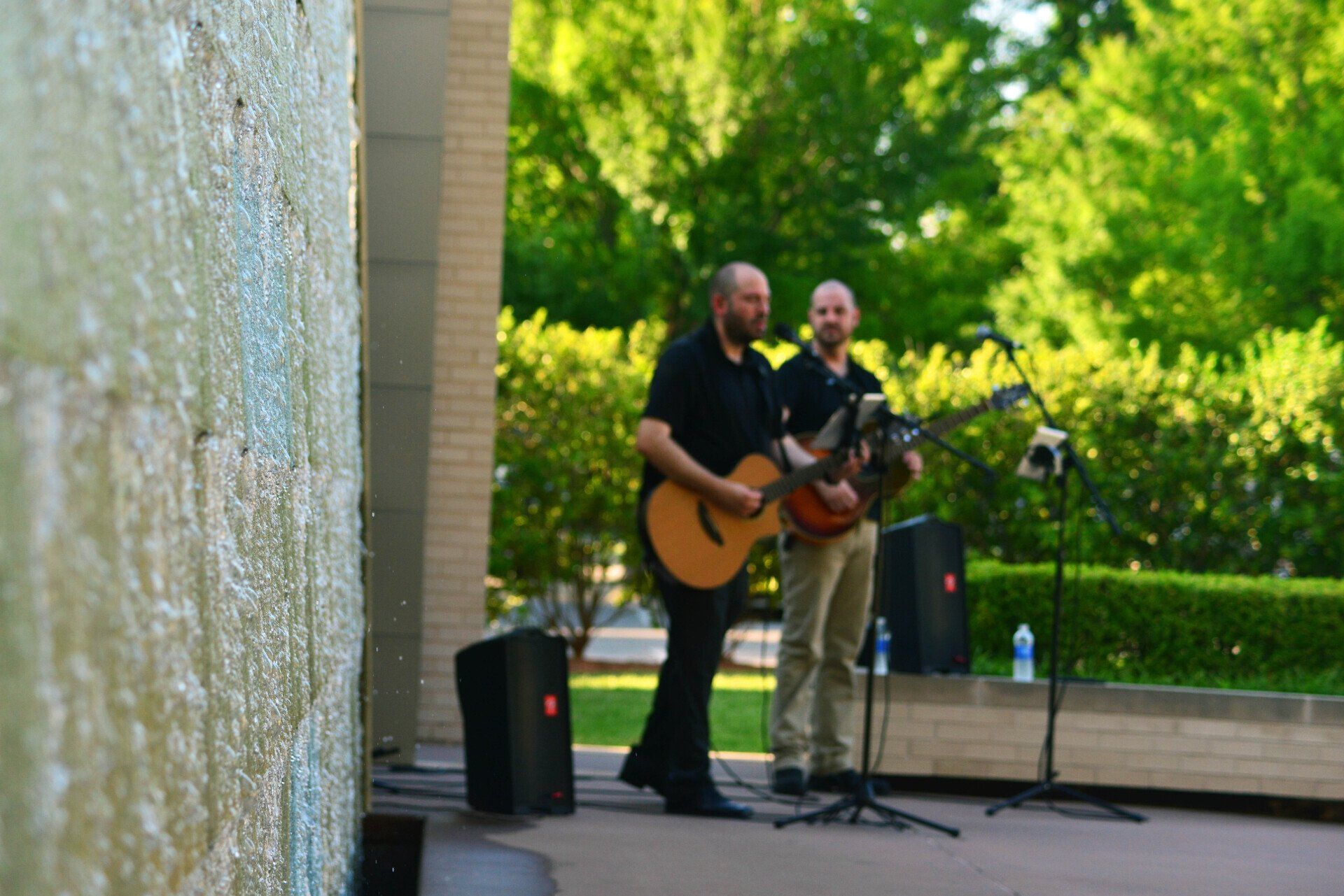 Two men playing guitars outdoors near a water feature and greenery. One man is singing and the other is playing.