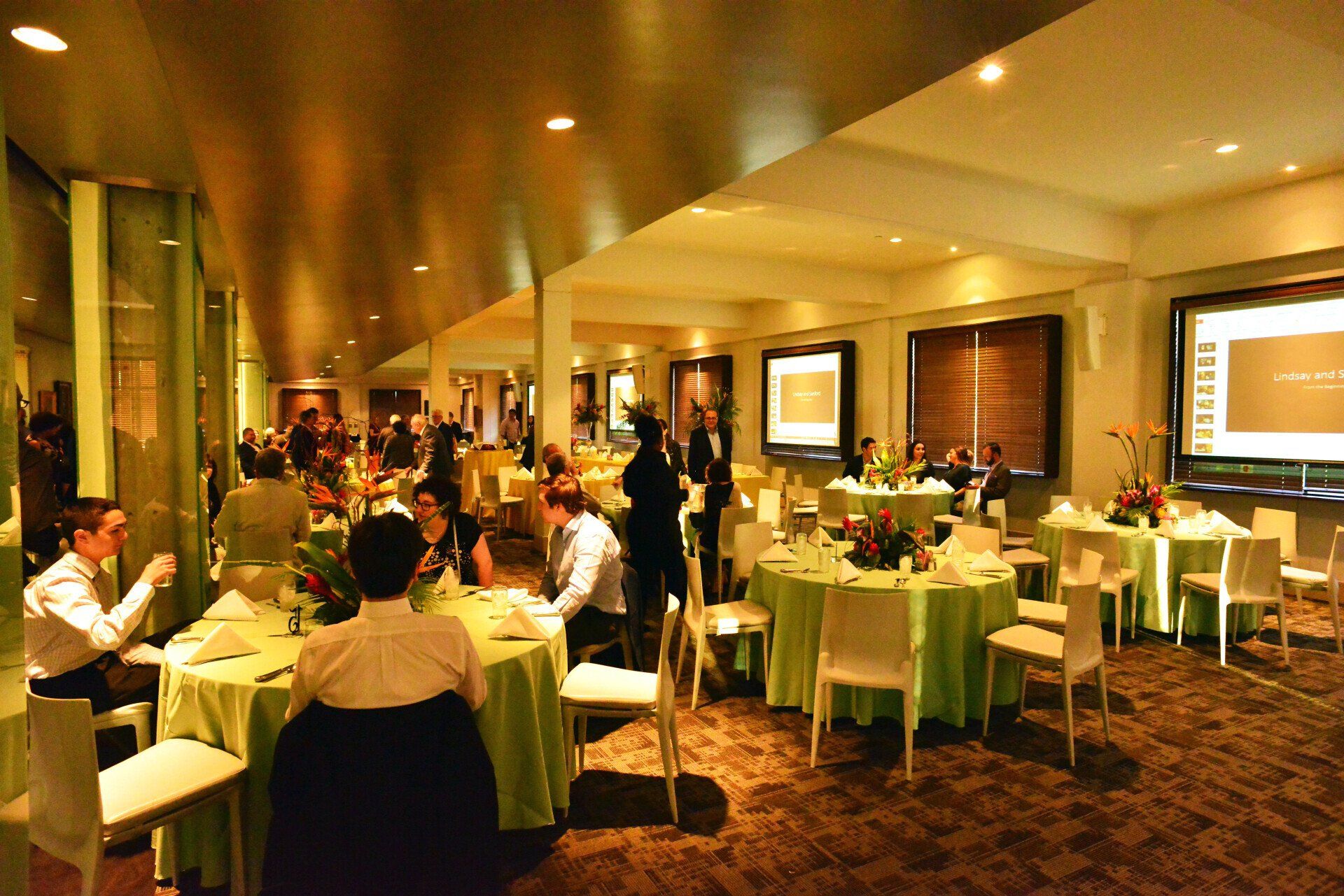 People gathered at round tables in a brightly lit banquet hall. Green tablecloths, white chairs, and artwork are visible.