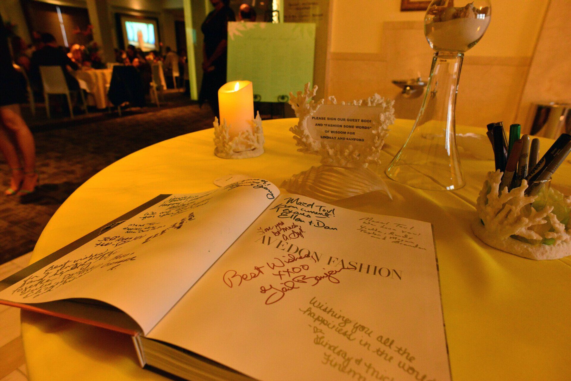 Open guest book on yellow table, with candle, pens, and decorative glass at an event.