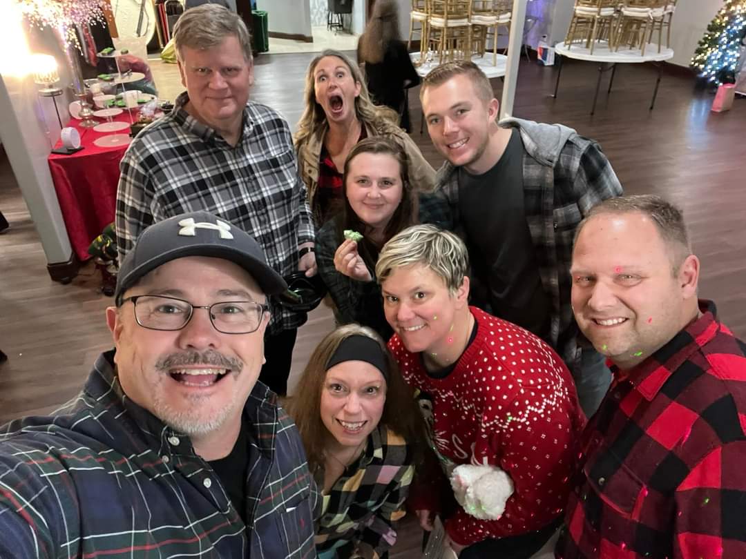 Group selfie in festive attire, indoors. People smiling, some with playful expressions. Christmas decorations in background.