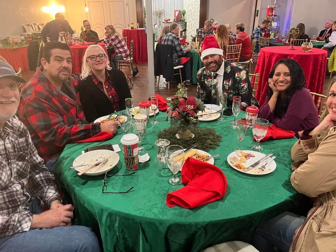 People at a festive Christmas party sit around a green table with red napkins and centerpieces.