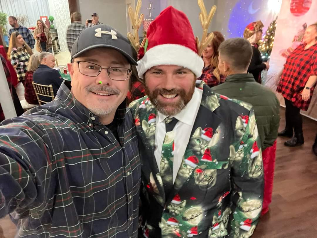 Two men smiling for a selfie at a Christmas party. One wears a festive cat suit and Santa hat.