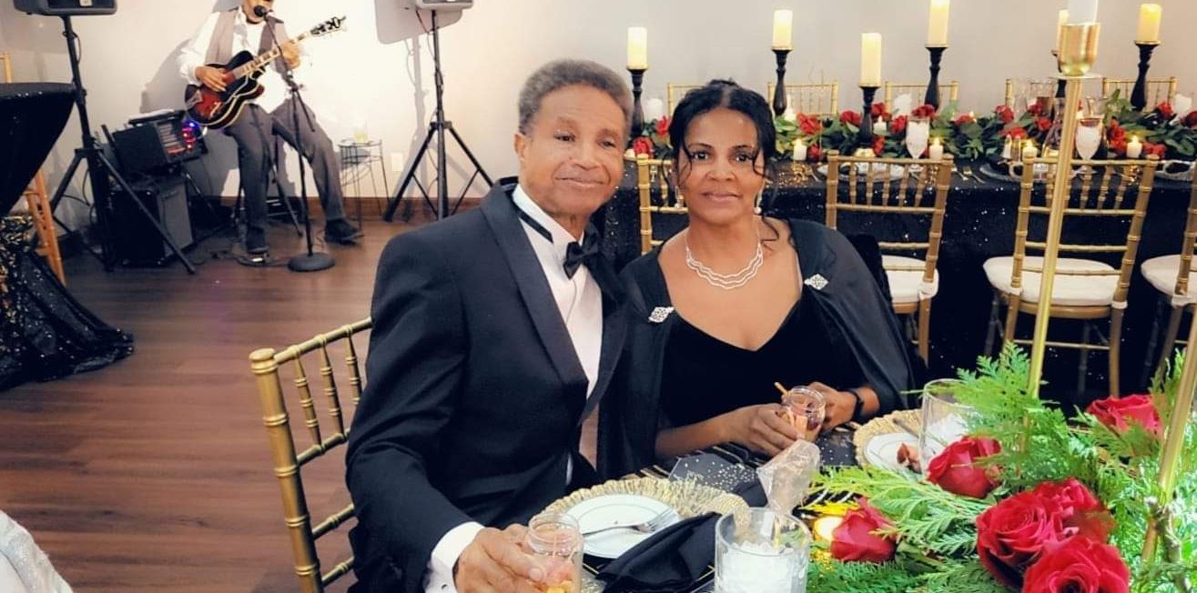 Couple at formal event; man in tuxedo, woman in black dress, candles and flowers on table.