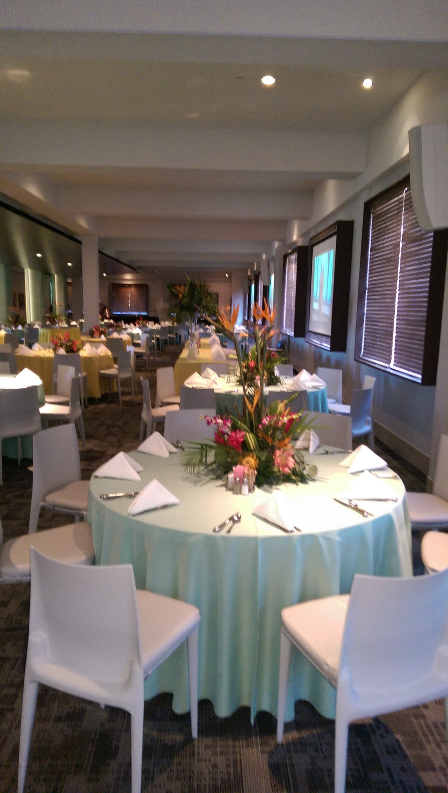 Round tables set for a banquet in a brightly lit room, with floral centerpieces and white chairs.