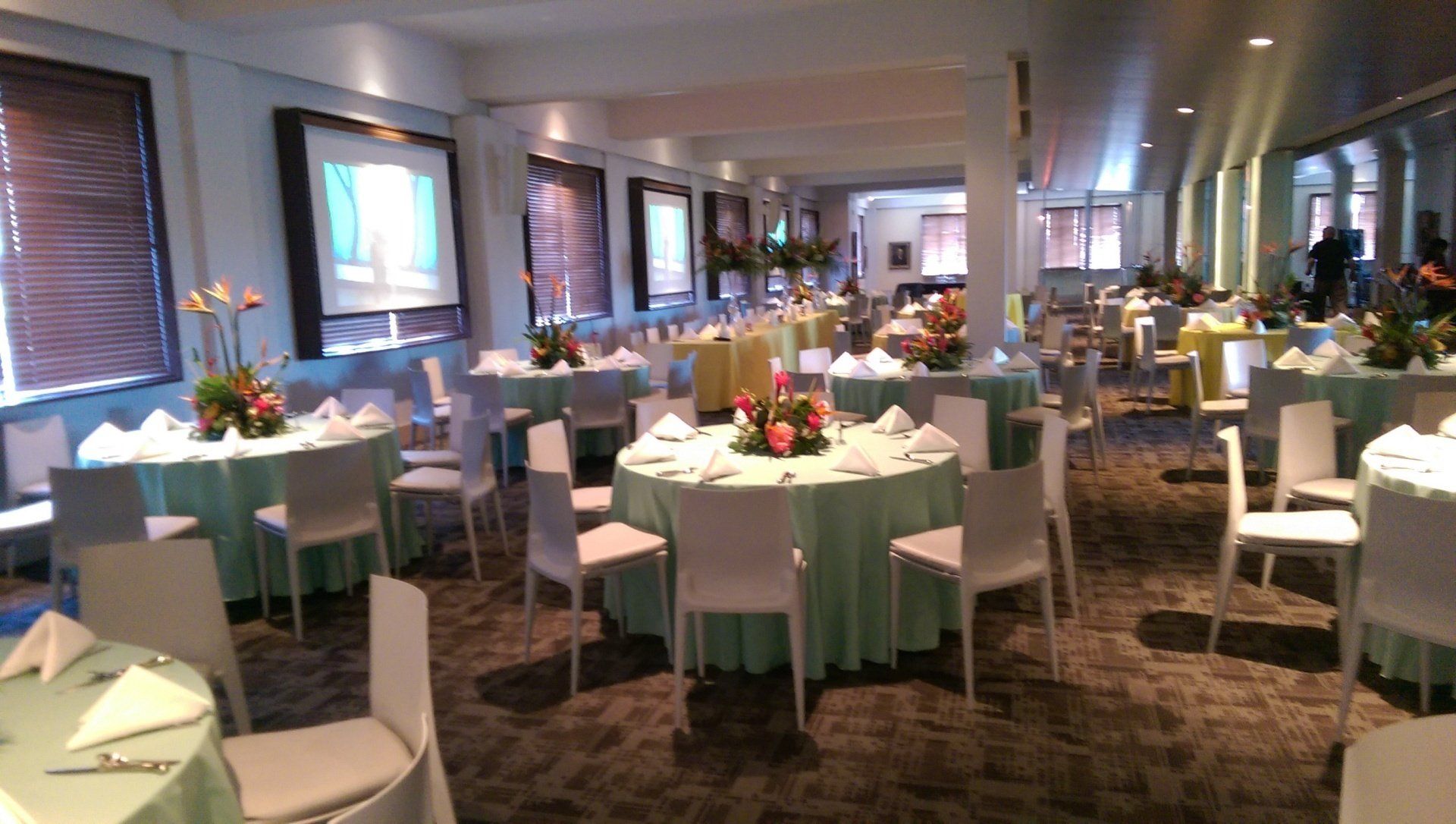 Round tables set for a banquet in a brightly lit room, each with a floral centerpiece.