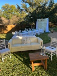 Outdoor event setup: white sofa, chairs, tables, and floral backdrop on grass.