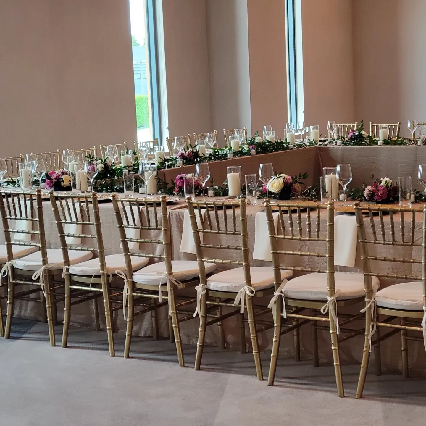 Long banquet table set for an event; gold chairs with white cushions, floral arrangements.