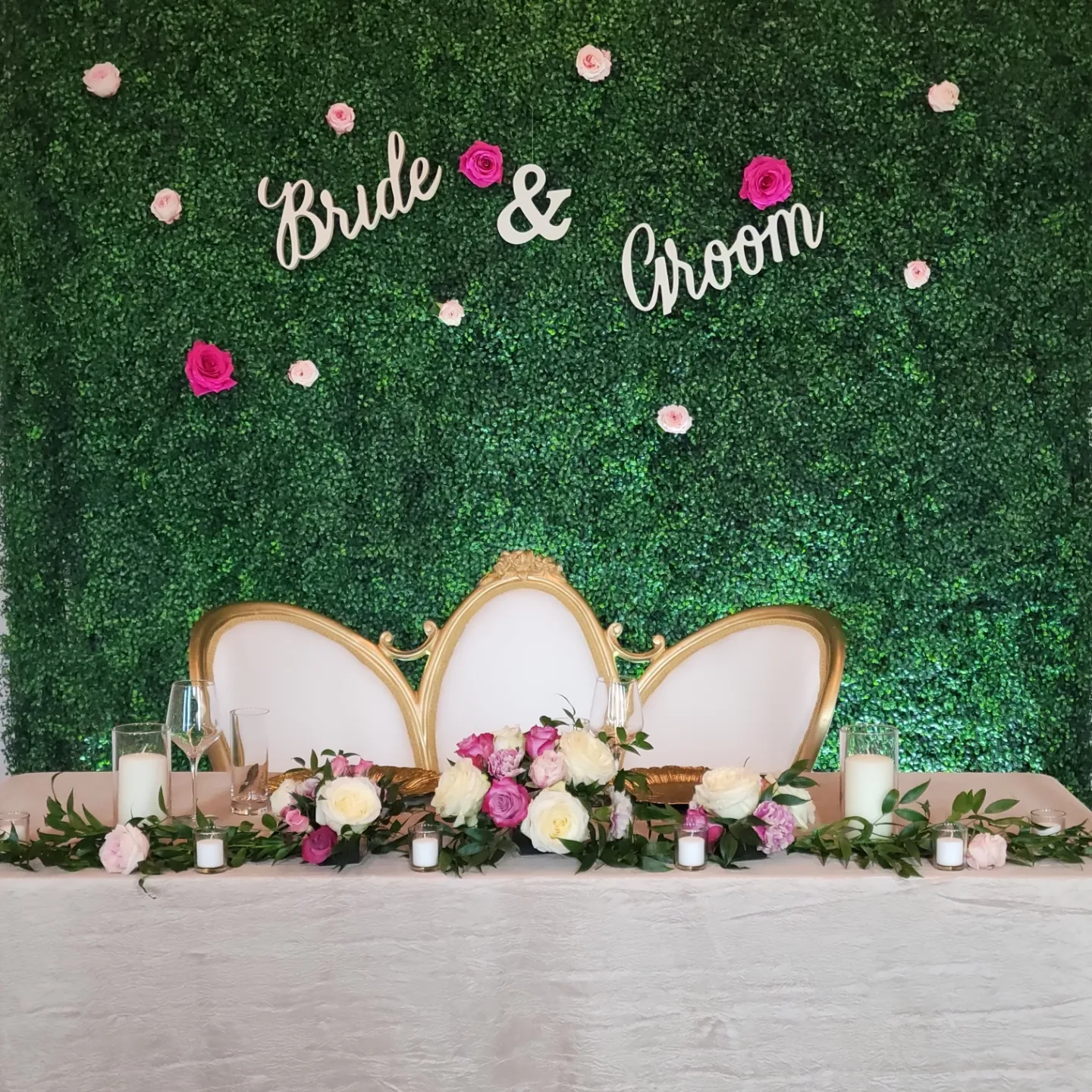 Bride and Groom table with floral arrangement, green backdrop, white and gold chairs.