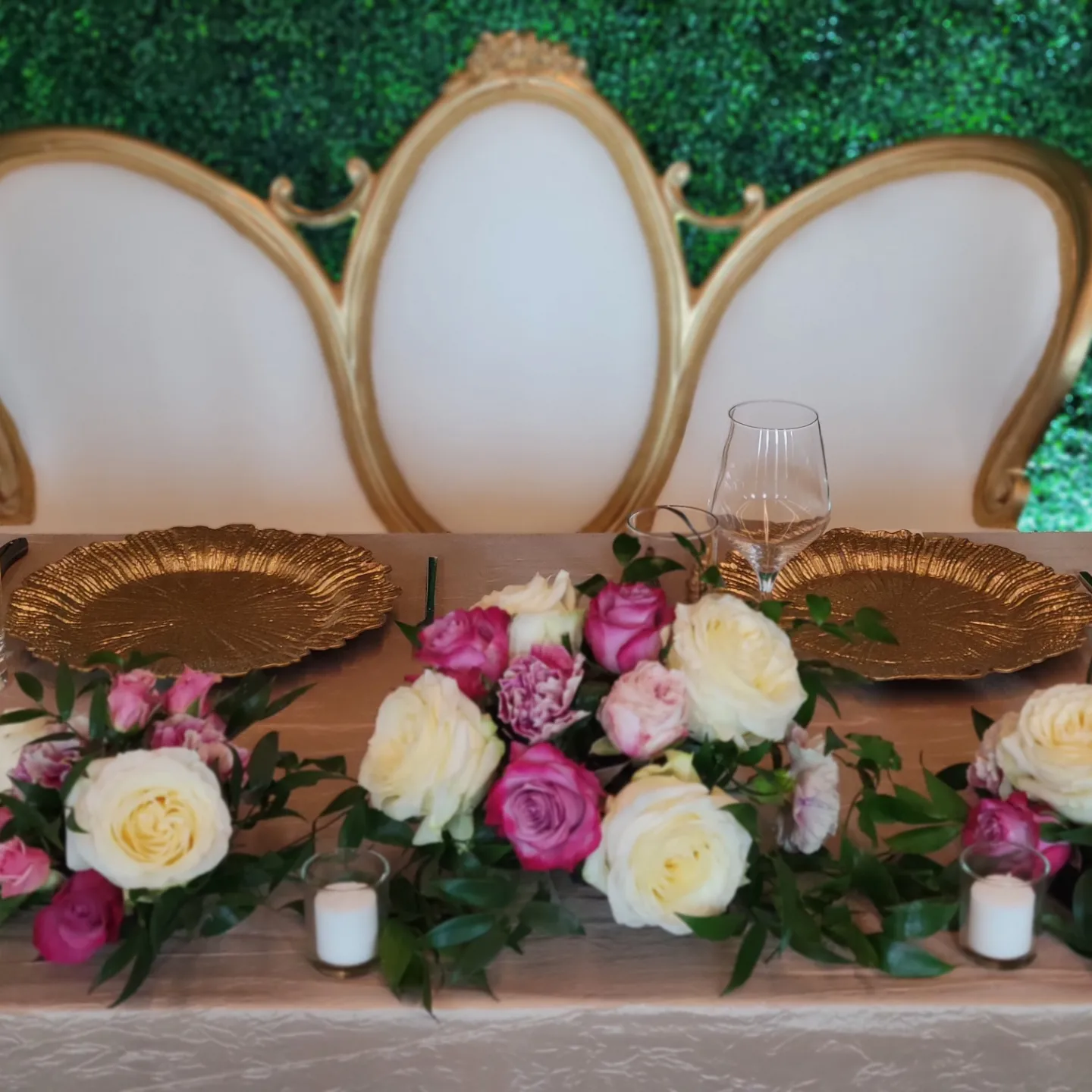 A sweetheart table set for a wedding with gold charger plates, floral arrangement, and white chairs.