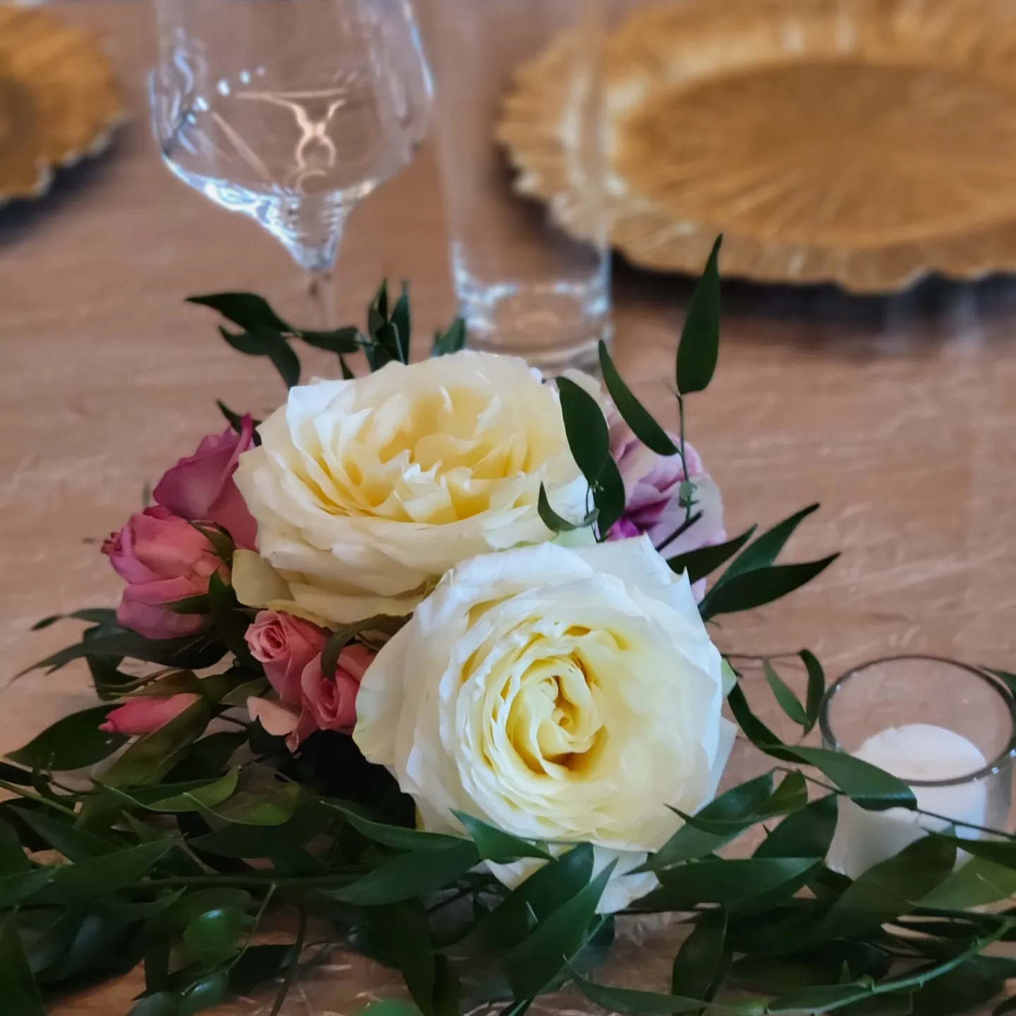 Floral centerpiece with white and pink roses, greenery, and a candle on a table.