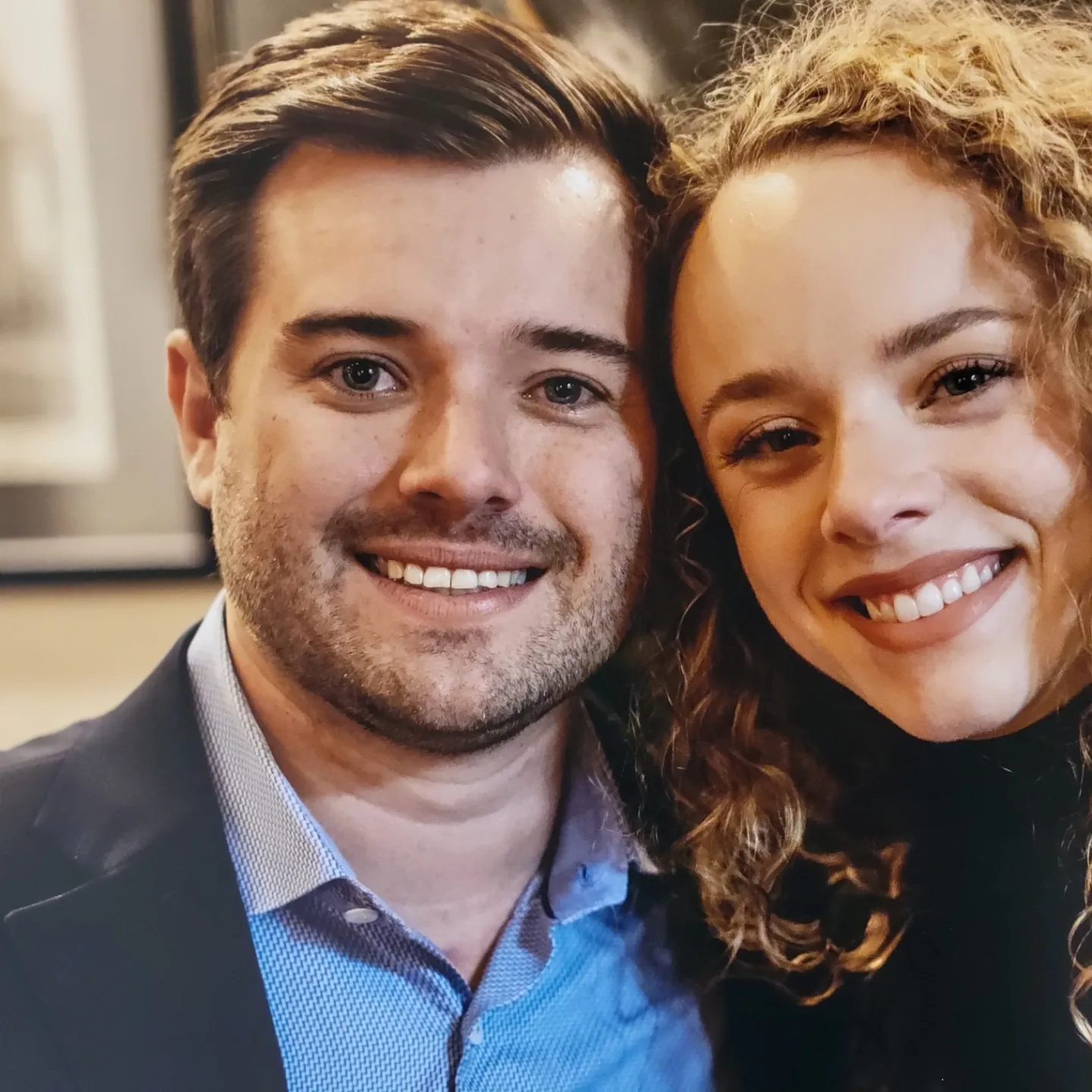 A smiling man and woman pose closely together. Man wears a blazer, woman has curly hair.