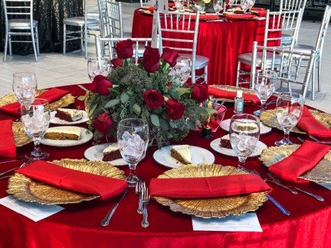 Red-themed event table setting with gold chargers, napkins, flowers, and cake slices. Glassware surround the table.