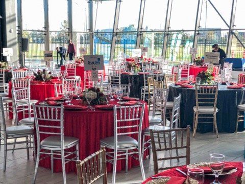 Tables set for an event with red, black, and blue tablecloths; clear and silver chairs; large windows.