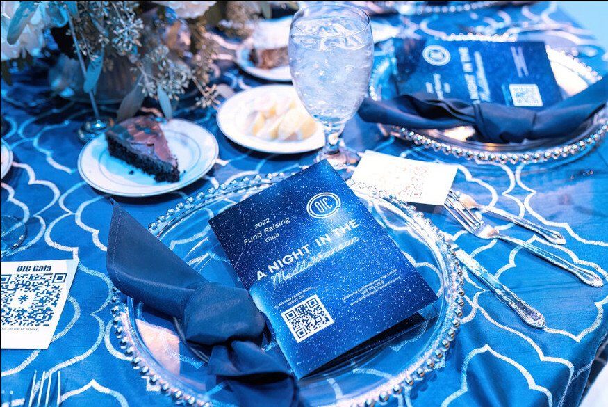 Table setting for an event. Blue tablecloth, silver charger plates, dark napkin, menu. Dessert plate and water glass present.