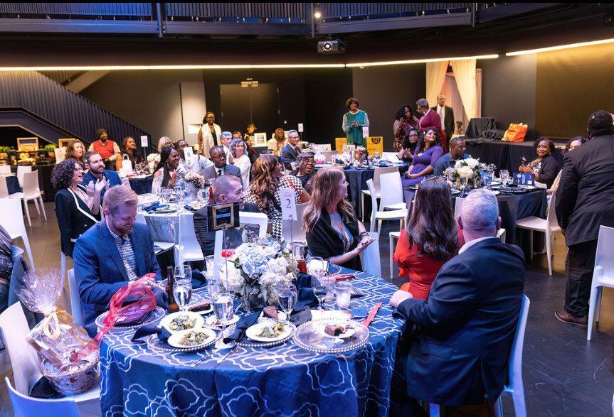 Formal event with seated guests at round tables. Blue tablecloths, white chairs, and floral centerpieces.