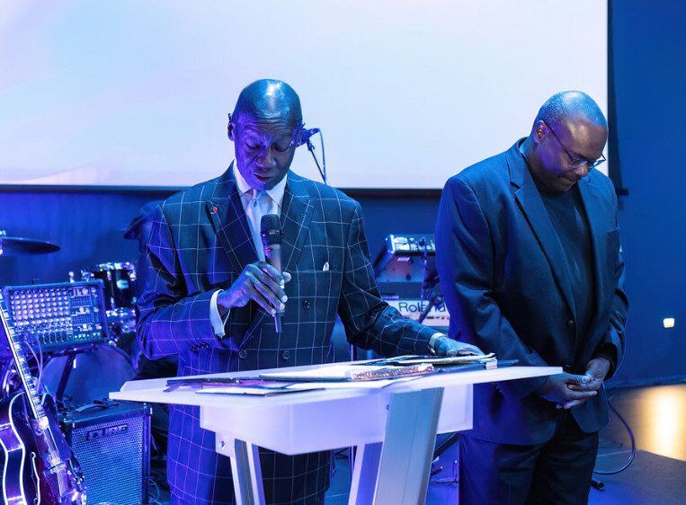 Two men at a podium; one speaks into a microphone, the other has hands clasped. Blue-lit stage.