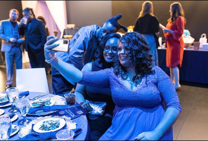 Three people pose for a selfie at a formal event, smiling. Blue lighting, people in formal attire.