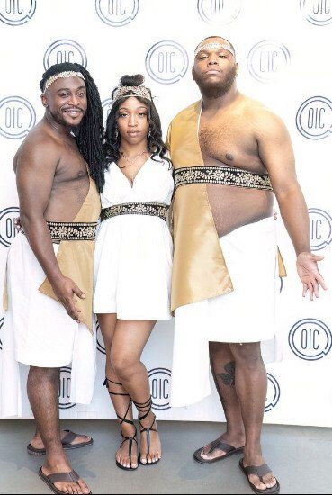 Three people in white and gold Roman-inspired costumes stand in front of a white backdrop with 