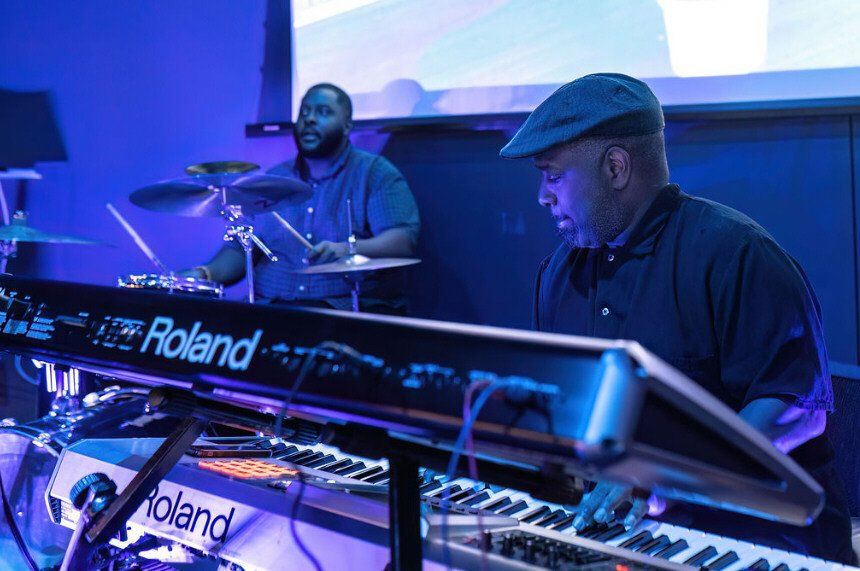 Two musicians performing on keyboards and drums under blue stage lighting.