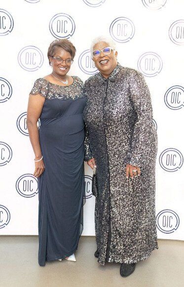 Two women pose in front of a backdrop with logos. One in a navy gown, the other in a sequined coat.