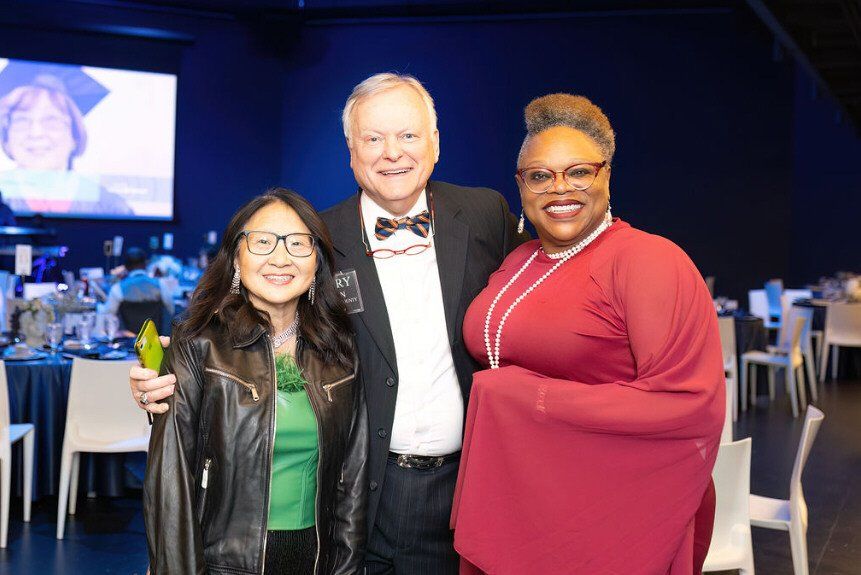 Three people smiling for a photo at an event; woman in red dress, man in bow tie, woman in leather jacket.