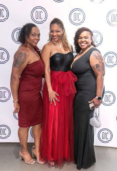 Three women in formal dresses pose at a black-tie event, smiling.