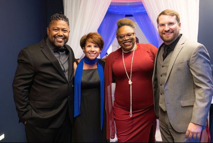 Four people smiling at an event; two men, two women, near a draped entryway.