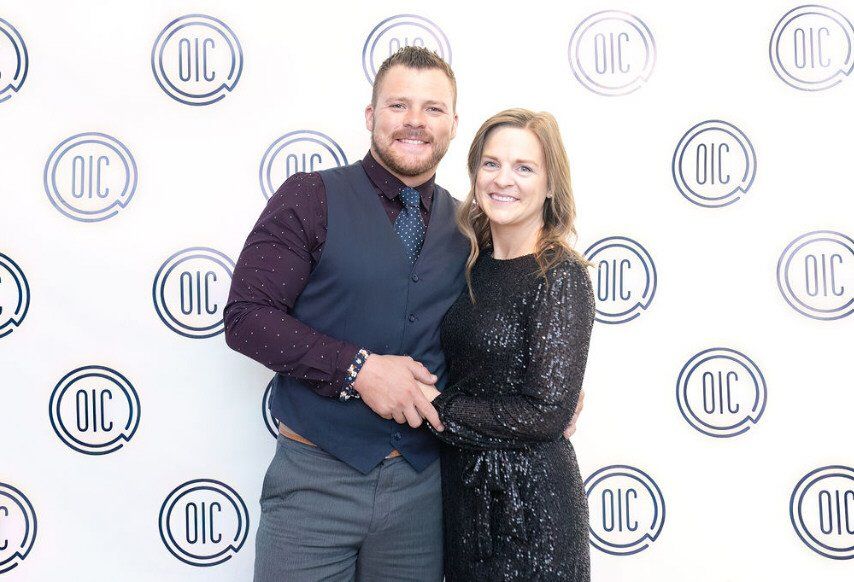 A man and woman pose in front of a backdrop with logos. The man wears a vest and the woman wears a sequined dress.