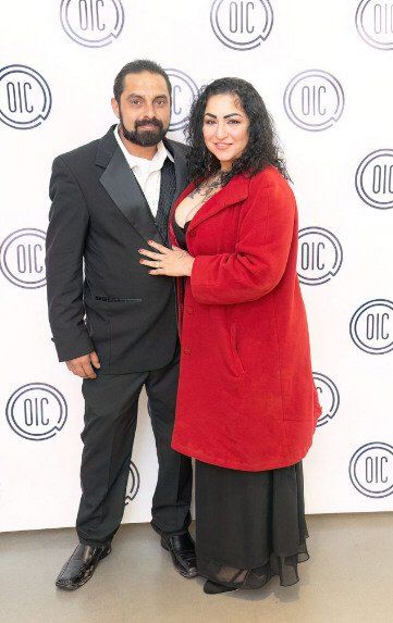 Couple posing at event, man in suit, woman in red coat, white backdrop with 