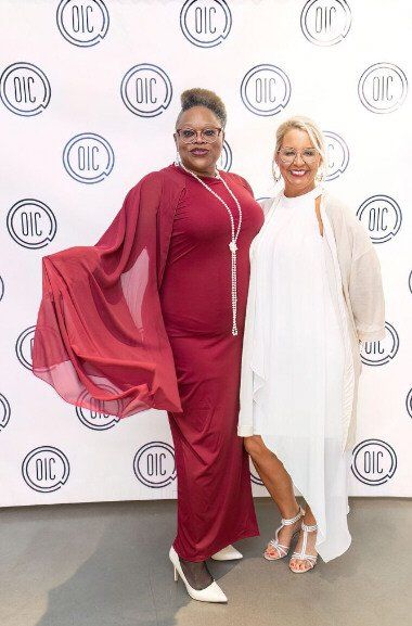 Two people pose in front of a backdrop with logos. One wears a red gown and heels, the other a white outfit and heels.