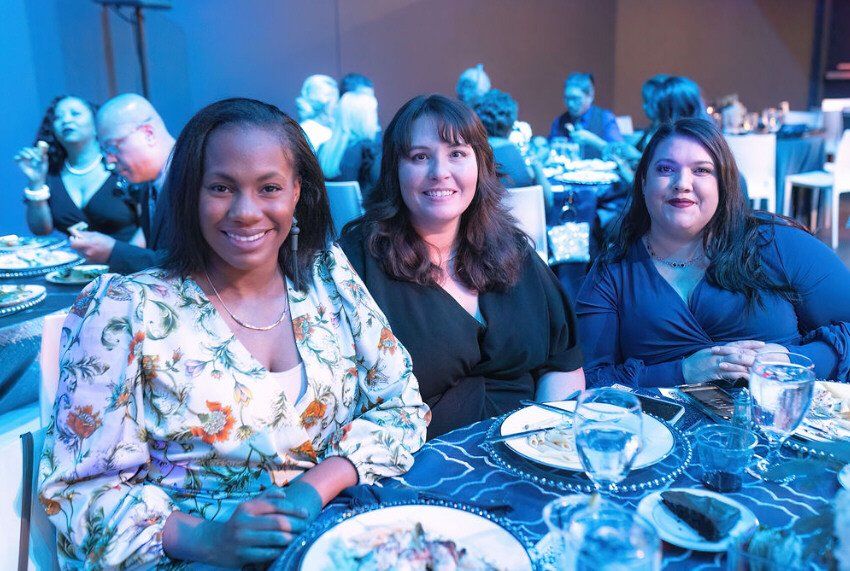 Three women smiling at a table set for dinner; indoor event with blue lighting.